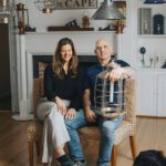 Chris and Kelly Berard of Nauset Lantern Shop seated in their Cape Cod studio surrounded by handcrafted lighting fixtures.