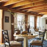 Cozy breakfast room with exposed beams, round wood table, and ladder-back chairs beneath a row of windows