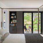 Minimalist bedroom with a modern four-poster bed, built-in shelving, and sliding glass doors opening to a wooded outdoor patio