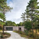 Modern Cape Cod home with flat rooflines and a gray facade, set along a wooded gravel driveway