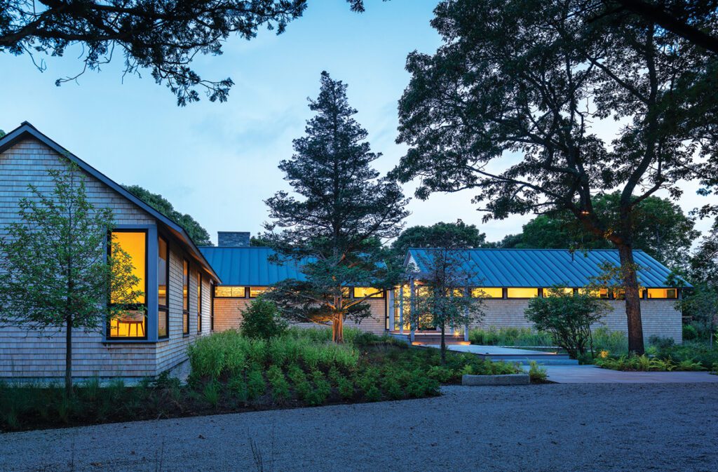 Twilight view of a modern shingle-style home with warm interior lighting and a striking linear design nestled into a wooded landscape.