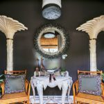 Dramatic black-walled hallway with an ornate white console, sunburst mirror, and statement palm leaf columns flanked by rattan chairs.