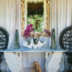 Decorative shell-encrusted mirror over a swan-base marble console table, flanked by black peacock chairs and topped with orchids in a clam shell bowl.