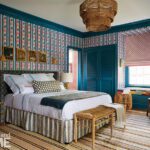 Eclectic bedroom with vibrant red, blue, and teal floral wallpaper, layered textiles, a woven chandelier, and a cozy window seat framed by painted trim.