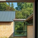 Contemporary coastal home featuring cedar shingles, standing-seam metal roof, and large corner window framed by natural landscaping.