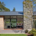 Modern home exterior with stone chimney, large glass doors, and shingled siding, showcasing indoor-outdoor connection and clean lines.