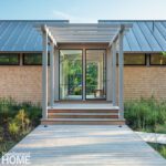 Front entry view of a minimalist shingle-style home with pergola and glass entryway, set against lush native plantings.