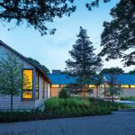 Twilight view of a modern shingle-style home with warm interior lighting and a striking linear design nestled into a wooded landscape.