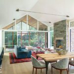 Open-concept living and dining room with floor-to-ceiling glass, stone fireplace, and natural wood finishes connecting to the outdoors.