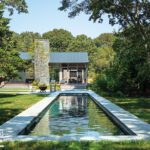 Long, narrow lap pool framed by natural landscaping and lawn, with a view toward the home’s modern glass and stone exterior.