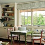 Cozy dining nook with green spindle-leg table, built-in bookcases, window bench seating, and scenic views through large double-hung windows.