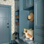 Charming mudroom with custom blue cabinetry, floral wallpaper, built-in bench, woven baskets, and a small dog sitting on a cushion.