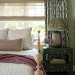 Serene bedroom with light wood bedframe, pink block-print pillows, floral drapery, and green nightstand topped with a woven lamp.