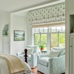 Bright bedroom sitting area with matching pale blue floral chairs, scenic window views, and delicate layered window treatments.