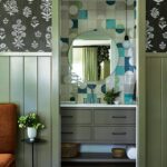 Peek into a bold bathroom with graphic tilework in shades of blue and green, a round mirror above a gray vanity, and floral wallpaper in the bedroom beyond.