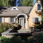 Charming stucco home with gray shutters, arched entryway, window boxes, and lush landscaping in a wooded New England setting.