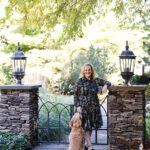 Pamela Foreman standing by a stone gate in a landscaped garden, smiling next to her dog under a tree canopy.
