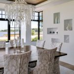 Elegant dining room with patterned upholstered chairs, a rustic wood table, and a sparkling crystal chandelier, offering water views through black-framed windows.