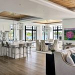 Open-concept living space with connected kitchen, dining, and sitting areas, featuring coffered ceilings and expansive water-facing windows.