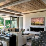 Inviting living room with layered textures, coffered wood ceiling, modern stone fireplace, and views of the water through large glass doors.