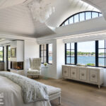 Light-filled primary bedroom with vaulted ceiling, custom chandelier, fireplace, and panoramic water views through black-trimmed windows.