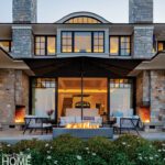 Elegant rear exterior at dusk with symmetrical stone chimneys, glowing interior lights, and a modern fire feature on the patio.