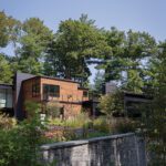 Modern hillside home with wood and metal facade surrounded by lush greenery and a terraced stone retaining wall.
