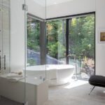 Luxurious bathroom featuring a freestanding soaking tub, glass shower, and floor-to-ceiling windows with wooded views.