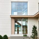 Modern home entrance with large glass windows and floating staircase visible inside.