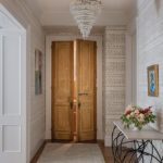 Hallway leading to primary suite with vintage wood doors, chandelier, and soft neutral wallpaper.