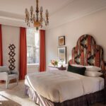 Guest bedroom with sculptural headboard, burnt-orange drapes, and vintage chandelier in warm natural light.