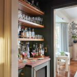 Elegant home bar with wood shelving, glassware, and dark green cabinetry designed by Erin Gates.