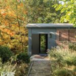 Brick and wood walkway leads to a minimalist gray and brick entrance surrounded by fall foliage.