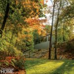 Modern structure with expansive windows perched on a rocky hillside surrounded by fall trees.