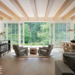 Open-concept living and dining space with exposed ceiling beams and floor-to-ceiling windows overlooking greenery.