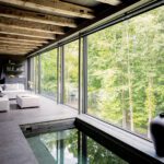 Sleek indoor lap pool with rustic exposed beams and full-height windows overlooking the woods.