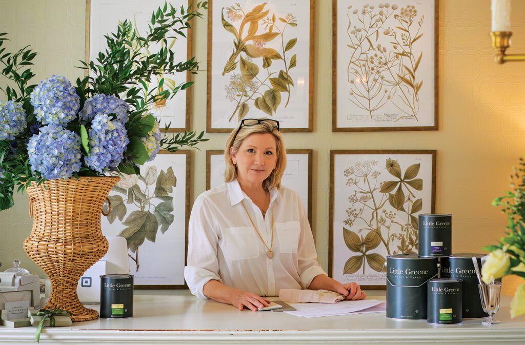 Designer Leslie-jon Vickory stands behind a white counter with floral arrangements and Little Greene paint cans, set against a backdrop of framed botanical prints and soft lighting.