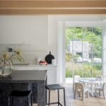Modern white kitchen with dark stone island and views to a garden through large grid windows.