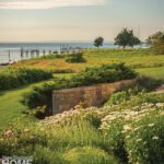 Coastal garden with stone retaining wall, lush greenery, and wildflowers overlooking a wooden pier and calm ocean waters.