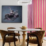 Dining area with round gold pedestal table, velvet chairs, and pink striped curtains.