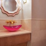 Glam powder room with pink glass vessel sink, gold fixtures, and scalloped mirror.