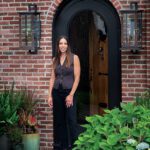 Interior designer Noelani Zervas stands at the entrance of a red brick home with black arched door