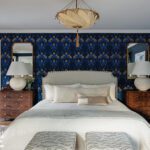 Deep blue primary bedroom with patterned wallpaper, upholstered headboard, and warm wood nightstands