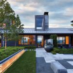 Modern home exterior with sleek metal roof, expansive windows, and lush landscaping at sunset.