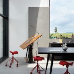 Modern home office with drafting table, red stools, and large windows showcasing meadow views.