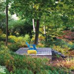 Modern deck with a blue chair and yellow ottoman nestled in a colorful woodland garden