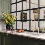 Kitchen sink with brass faucet, marble counters, and black grid window.
