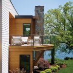 Side view of modern lake house with wood siding, balcony, and stone chimney.
