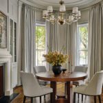 Dining room with round wood table, gray upholstered chairs, and chandelier in a bay-window alcove.