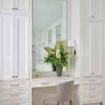 White built-in dressing table with marble floor, tall cabinetry, and mirror accented by fresh flowers.